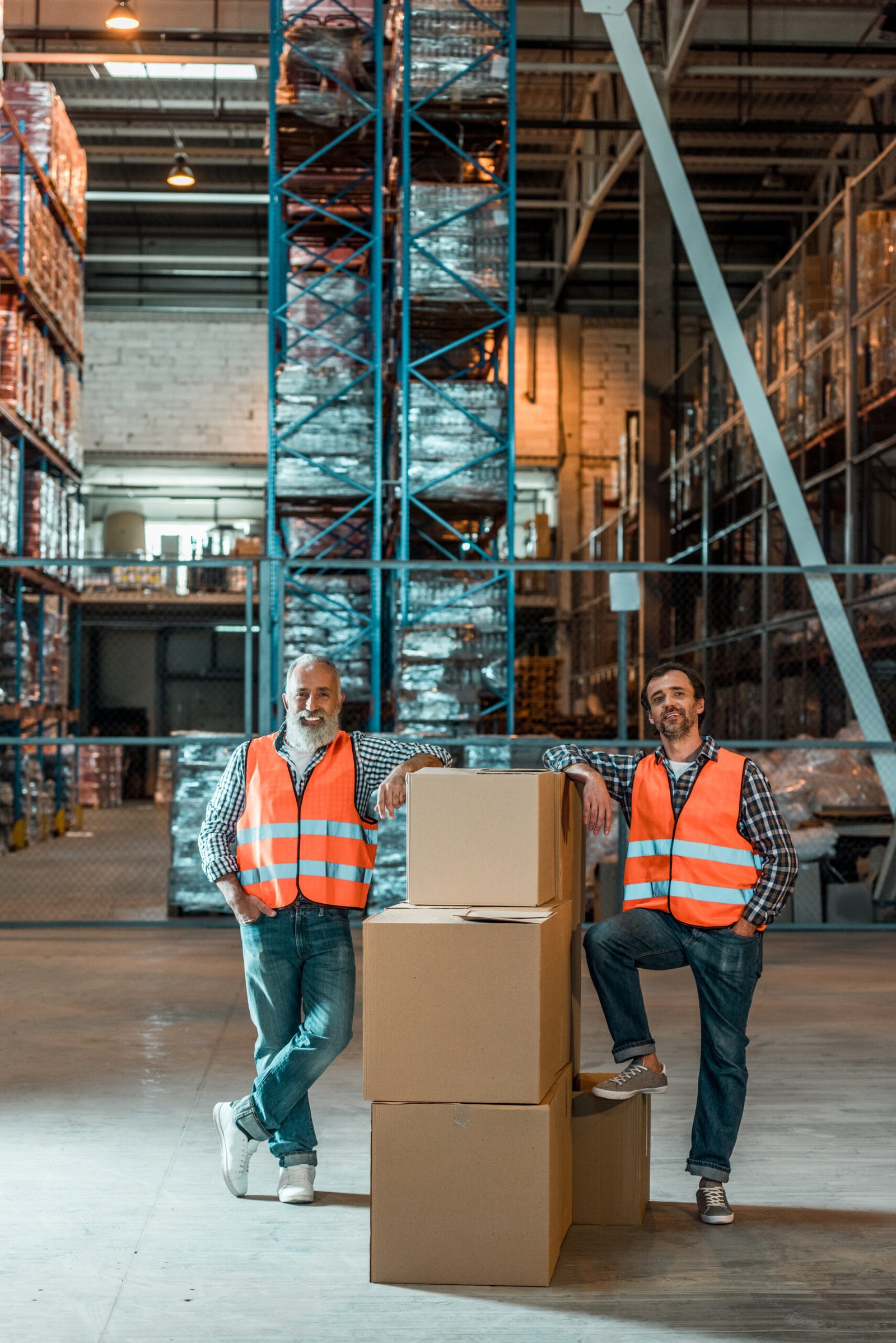 male warehouse workers in vests standing with boxes and smiling at camera